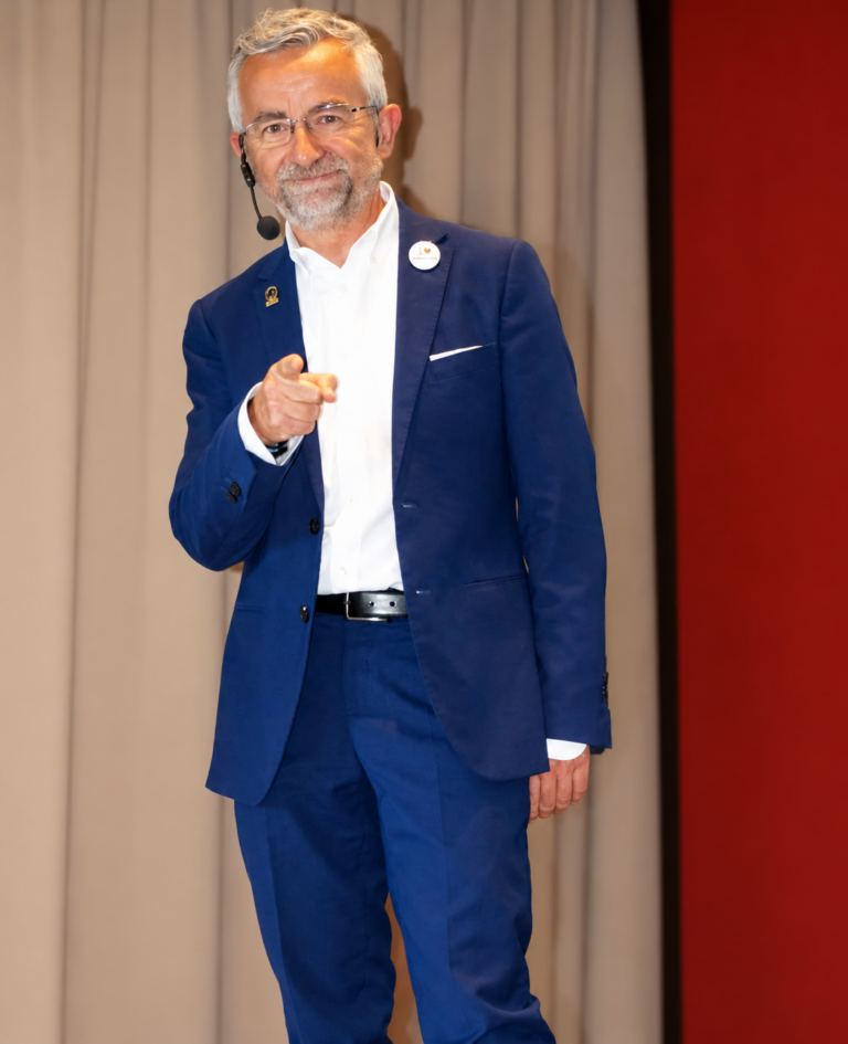 Fabio Gelmetti in abito blu con microfono ad archetto durante un intervento pubblico.